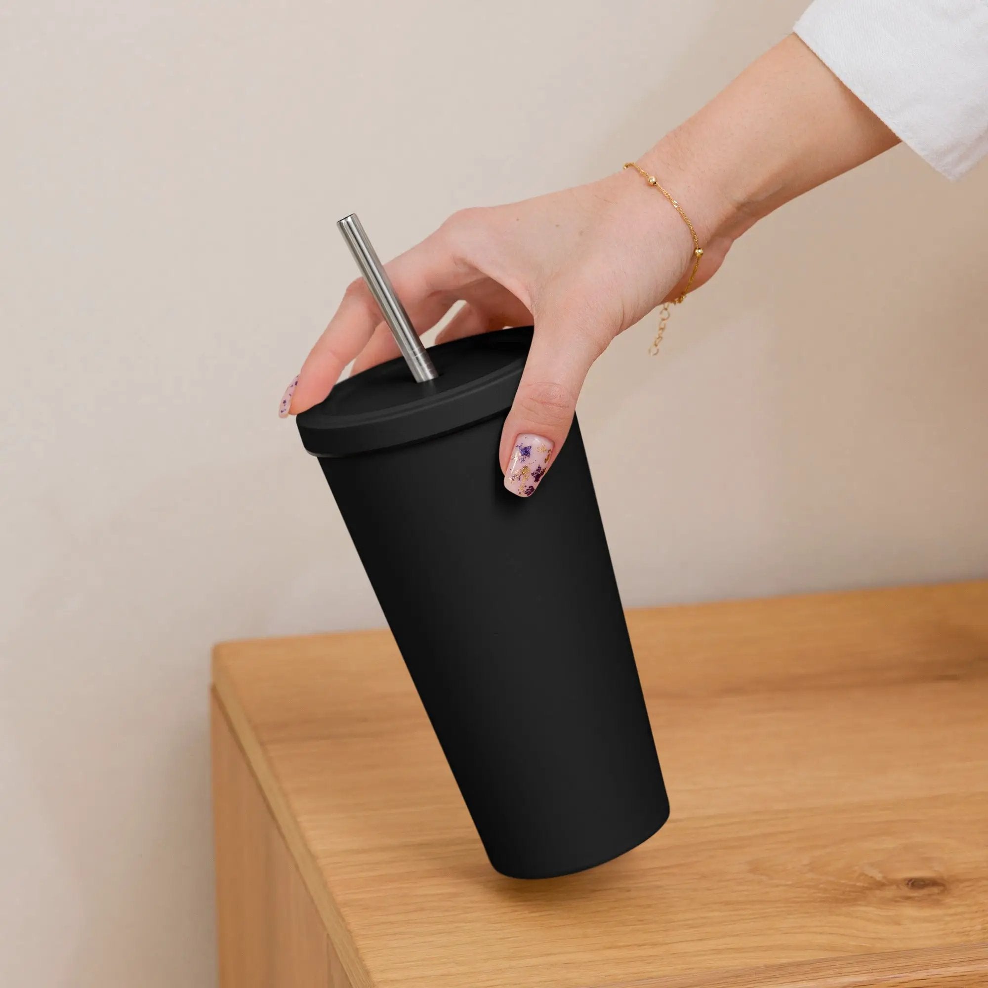 Insulated tumbler with metal straw in a black matte finish, held by a hand with nail art and a gold bracelet.