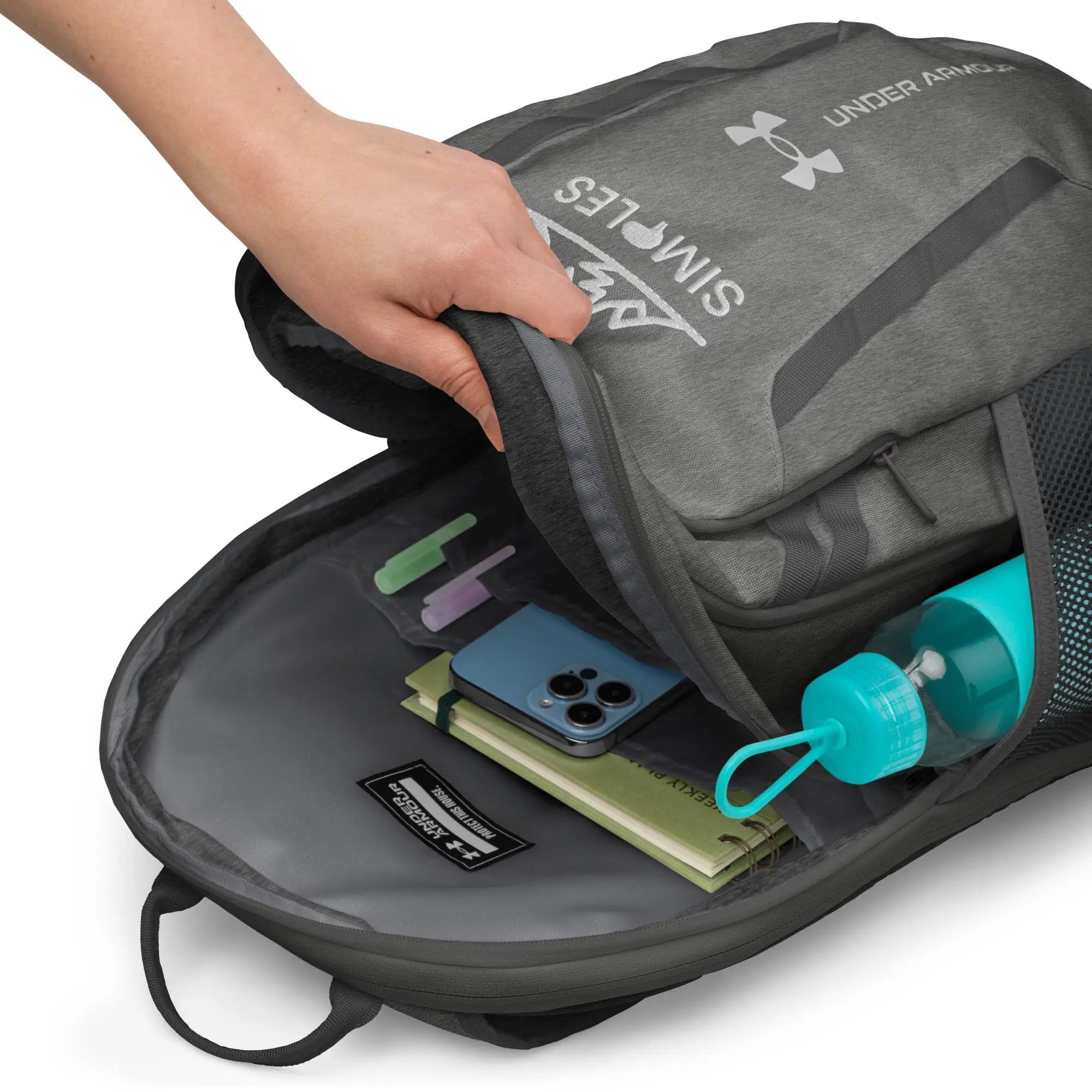 Under Armour backpack, gray fabric with logo, open main pocket showing phone, notebook and water bottle.